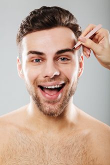Portrait of joyful man smiling while female hand plucking his eyebrows with tweezers, isolated over gray background
